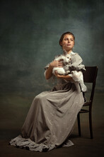 Portrait Of Young Beautiful Girl In Gray Dress Of Medieval Style Sitting With Fluffy Rabbit Isolated On Dark Background.