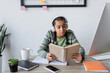 © LIGHTFIELD STUDIOS - african american teenage girl in headphones reading book near computer and smartphone with blank screen