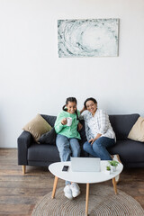 Wall Mural - smiling african american girl with cup of tea watching movie on laptop together with mom