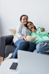 Wall Mural - smiling african american woman with popcorn watching movie on blurred laptop with amazed daughter