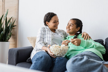 Wall Mural - happy african american woman with bowl of popcorn sitting on sofa with teenage daughter