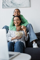 Wall Mural - smiling african american girl embracing laughing mother holding bowl of popcorn while watching film on blurred laptop