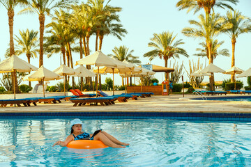 Wall Mural - A woman in a sun hat in the pool in a swimming circle with a laptop works remotely on the background of sun loungers.