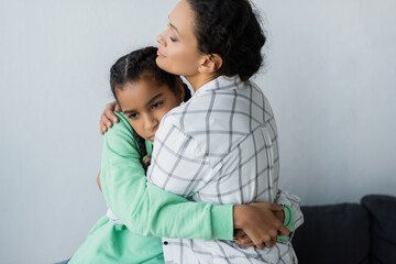 Wall Mural - african american mother with closed eyes embracing depressed teenage daughter at home