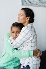 Wall Mural - african american woman with closed eyes embracing frustrated daughter at home