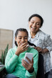 © LIGHTFIELD STUDIOS - cheerful african american woman braiding hair of laughing daughter looking at mobile phone