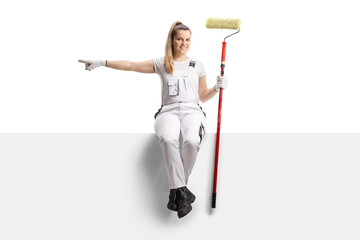 Sticker - Full length portrait of a female house painter sitting on a blank panel with a paint roller and pointing to the side