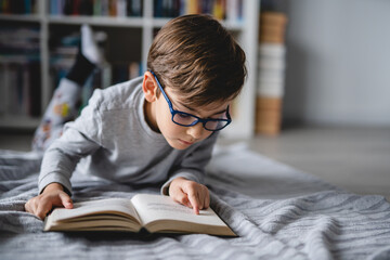 Wall Mural - One caucasian boy lying on the floor at home in day reading a book front view copy space real people education concept