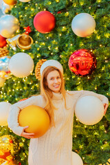 Wall Mural - A smiling girl holds two huge Christmas balls under her arms against the backdrop of a giant elegant fir tree. Festive New Year atmosphere.