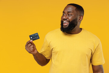 Wall Mural - Young smiling satisfied fun rich cool happy black man 20s wearing bright casual t-shirt hold in hand credit bank card look aside on workspace isolated on plain yellow color background studio portrait.