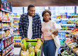 © Prostock-studio - Portrait of happy black couple with shopping cart full of products choosing food together at modern supermarket