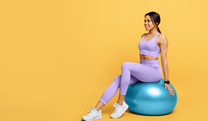 Wall Mural - Fitness exercises. Happy african american lady sitting on fitball and looking at empty space over yellow background