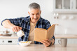 Happy senior man reading book and poring milk in cereals, having breakfast in kitchen interior