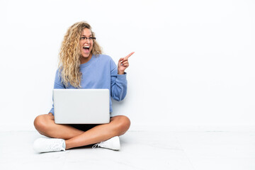 Wall Mural - Girl with curly hair with a laptop sitting on the floor pointing finger to the side and presenting a product