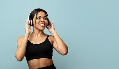 Wall Mural - Active free time. Sporty black lady in sports uniform with headphones exercising or dancing, blue background, free space