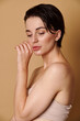 © Taras Grebinets - Confident attractive woman with healthy glowing skin in beige underwear looking down, touching her face, posing against beige colored background with copy space. Beauty treatment, cosmetology concept