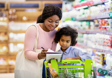 Millennial black mother and her adorable daughter checking grocery list in notebook while shopping at supermarket