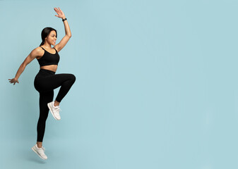 Wall Mural - Fitness concept. Fit young african american woman jumping, lifting leg up, blue background with empty space