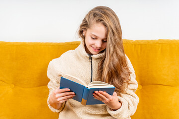 A little teenage girl is sitting on a yellow sofa at home and reading a book