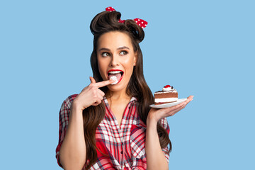 Wall Mural - Sexy young pinup woman in retro style outfit eating piece of yummy cake, licking her finger on blue studio background