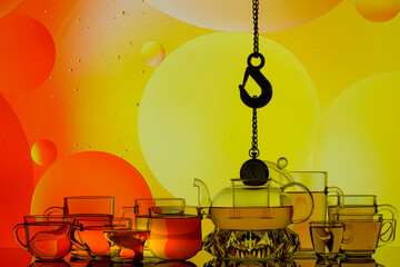 Wall Mural - transparent glass teapot and cups with tea and drops, silhouette of lifting hook on colorful background 