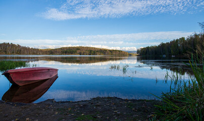 Naklejka na meble A red old wooden boat anchored near the shore, in a secluded harbor.
Landscape photography of a beautiful blue lake and an anchored red boat.