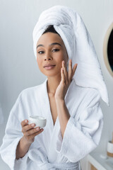 Wall Mural - african american woman in bathrobe holding container and applying face cream in bathroom