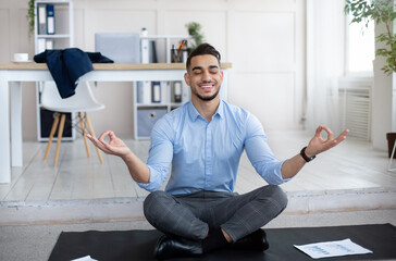 Wall Mural - Workplace stress management concept. Calm Arab man meditating with closed eyes, doing yoga at modern office