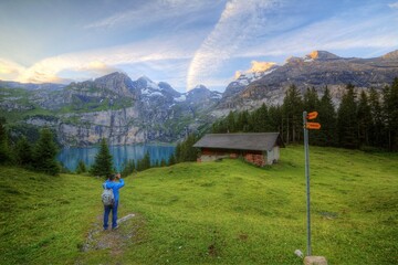 Wall Mural - Tourist enjoying the scenery of peaceful Oeschinen Lake with a wooden farm house by the lakeside & majestic mountains in background on a beautiful summer morning in Bernese Oberland, Switzerland