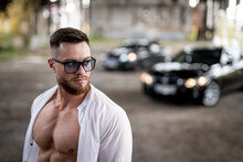 Handsome Man Is Standing In Unbuttoned White Shirt Near Broken Concrete Wall. Abandoned Storehouse And Car On The Background