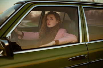 Fashionable confident woman posing at the wheel of green retro car. Elegant redhead model wearing classic pink suit. Copy, empty space for text 