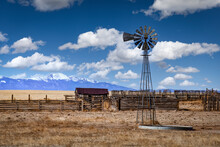 Old Farm With A Windmill During The Day With Fluffy White Clouds In The Sky. The Ground Is Brown And Barren. The Snow Capped Rocky Mountains Are Visible In The Distance. 