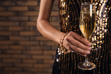 Woman In Beautiful Dress Holding Glass With Champagne Against Brick Wall, Closeup