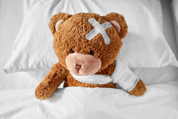 Wall Mural - medicine, healthcare and childhood concept - ill teddy bear toy with bandaged paw and medical patch on head lying in bed