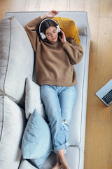 Wall Mural - Beautiful woman listening to music with headphone while using smartphone lying on sofa at home.