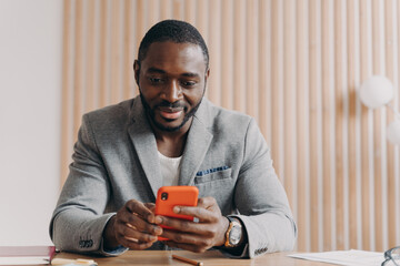 Young smiling african male entrepreneur sitting at office desk and using mobile phone at workplace