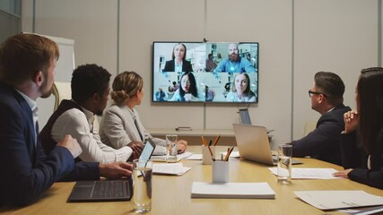 Dolly-in shot of team of businesspeople sitting at table in meeting room and discussing work with their colleagues via video call