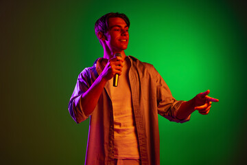 Wall Mural - Portrait of young man, pop singer with microphone singing isolated on green studio background in neon light