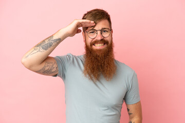 Young reddish caucasian man isolated on pink background saluting with hand with happy expression