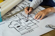 © Nut - Young engineer holding a black pencil pointing to a building on blueprints for a workplace meeting, hands of an engineer working with tools on a background on a project sketch.