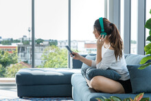 Asian Young Female Teenager Model In Casual Outfit Sitting On Cozy Sofa Couch With Pillows Wearing Headphones Listening To Music Changing Track Song Playlist From Smartphone In Living Room At Home