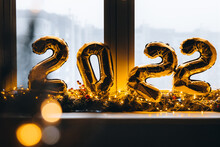 2022 Golden Foil Balloons On Blue Window Sill. Celebrating Holidays At Home, Festive Decor Concept. Happy New Year 2022. Close-up Numbers Of Year 2022 On Dark Background. Bokeh Warm Garland Light.