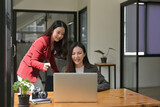 Fototapeta Tulipany - Photo of two beautiful office girls working together at the wooden working desk surrounded by a computer laptop, potted plant, camera and notebook.