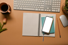 Top View Image Of A Cozy Workspace Surrounded By A Notebook, Keyboard, Pencil, Potted Plant, Wireless Mouse, Coffee Cup, Tulip Flower And White Blank Screen Smartphone.
