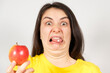A woman with a disgruntled grimace of pills on her tongue holds an apple. Rejection of synthetic dietary supplements and vitamins.