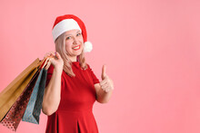 A Woman In A Santa Hat With Gift Bags On A Pink Background Shows A Thumbs Up Gesture