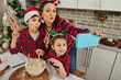 © Taras Grebinets - Cheerful beautiful Hispanic mom with antler hoop with her adorable children in elf hoop and Santa hat having fun together, cooking Christmas bread and making selfie looking at web camera in the phone