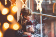© Irina Schmidt - Little preschool girl holding cup Santa Claus boot with gift called Nikolausstiefel in German. Happy child wait on holiday by window with Christmas lights in winter. Cozy family celebration of xmas.