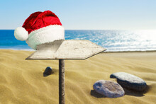 Wooden Sign Wearing A Santa Claus Hat, Against The Backdrop Of Sand Dunes And The Ocean. Close-up. Copy Space. Place For Your Text. New Year Card.