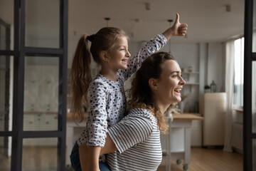 Happy mother piggy backing little daughter showing thumb up, dreaming of good future, excited loving young mom and adorable 8s girl child engaged in funny activity, family having fun at home